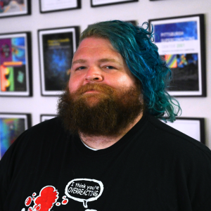 A person with light skin tone, a beard, and blue hair smiles at the camera.